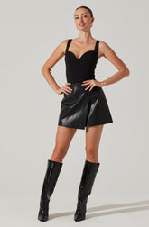 The woman is wearing a fitted black sleeveless top with wide shoulder straps and a sweetheart neckline. She pairs it with a high-waisted black leather mini skirt with a front slit. She completes her outfit with knee-high black patent leather boots.